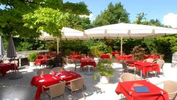 Außenansicht der Terrasse mit roten Tischdecken, umgeben von Bäumen und Sonnenschirmen.