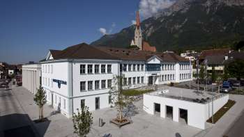 Das Domus-Gebäude in Schaan mit weißer Fassade und Blick auf die Pfarrkirche – ein kulturelles Zentrum und Begegnungsort im Herzen von Liechtenstein