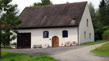 Traditionelle Mühle in Eschen mit weißer Fassade, dunklen Fensterläden und altem Mühlstein-Ensemble vor dem Eingang – umgeben von grüner Wiese und Bäumen.