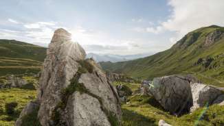 Sonnenaufgang hinter Felsformationen im grünen Naaftal in Liechtenstein, umgeben von sanften Hügeln und Berglandschaft
