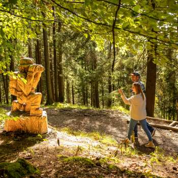 Frau zeigt auf eine detailreiche Holzskulptur im Wald am WalserSagenWeg