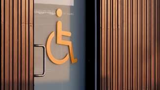 Orangefarbenes Rollstuhl-Symbol auf einer Glastür, das auf eine barrierefreie Toilette hinweist, mit Holzverkleidung im Hintergrund.