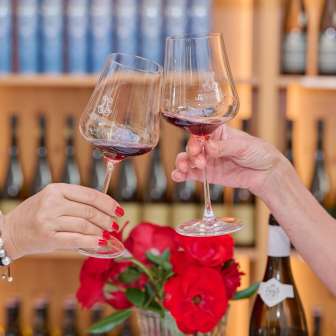 Zwei Frauen stoßen mit Rotwein in der Hofkellerei Liechtenstein an, im Hintergrund Weinregale mit edlen Tropfen