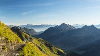 Alpiner Wanderweg mit Blick auf eine grüne Berglandschaft und schroffe Gipfel in der Morgensonne