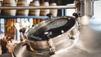 Nahaufnahme eines runden Sichtfensters an einem glänzenden Braukessel mit Blick auf die Sudhalle im Hintergrund.