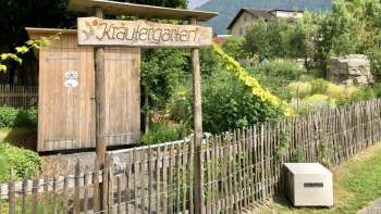 Holzschild mit Aufschrift „Kräutergarten“ am Eingang des liebevoll angelegten Gartens in Mauren, umgeben von Natur und Bergen.