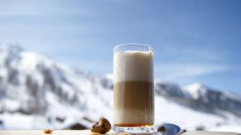 Glas Latte Macchiato auf einer Holzplatte vor der Kulisse verschneiter Berge