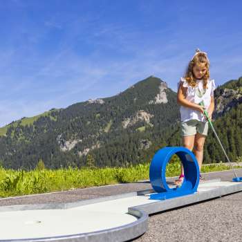 Mädchen konzentriert sich beim Minigolfspielen auf eine blaue Looping-Bahn in alpiner Kulisse
