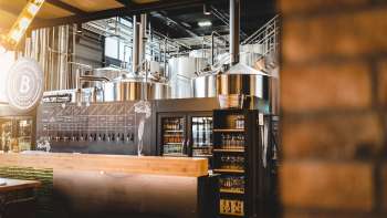 Innenansicht einer Brauerei mit Brauanlage aus Edelstahl und einladendem Schanktresen im Vordergrund.