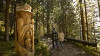 Zwei Wanderer betrachten geschnitzte Holzfiguren am WalserSagenWeg, einem Themenweg inmitten alpiner Landschaft mit Blick auf die Berge