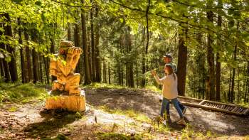 Frau zeigt auf eine detailreiche Holzskulptur im Wald am WalserSagenWeg
