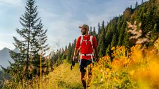 Wanderer mit rotem Shirt auf einem sonnigen Bergweg bei Malbun, umgeben von Wiesenblumen, Nadelwald und alpiner Kulisse in Liechtenstein