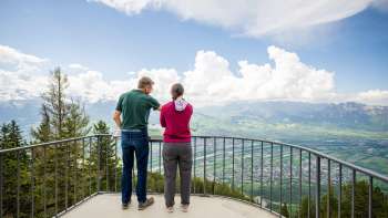 Zwei Personen genießen die Aussicht vom Aussichtspunkt Gaflei über das Rheintal in Liechtenstein bei klarem Wetter.