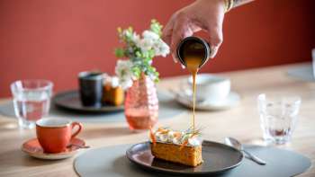Dessert wird mit Karamellsauce übergossen, im Hintergrund Kaffeetassen und stilvolle Tischdeko