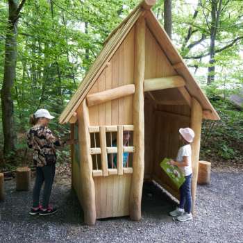 Zwei Kinder erkunden eine kleine Holzhütte im Wald, die Teil des Erlebniswegs „Wurz und Zirp auf Zeitreise“ in Liechtenstein ist – ein Naturabenteuer zum Anfassen.