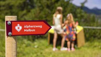 Wegweiser des Alpherzwegs Malbun mit Kindern vor einer der Stationen im Hintergrund
