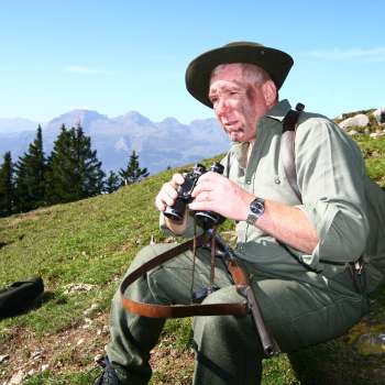 Ein Jäger mit Fernglas sitzt auf einer Wiese vor alpiner Bergkulisse.