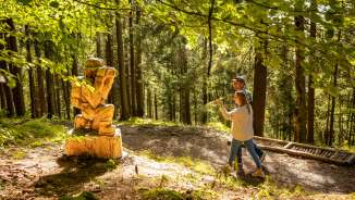 Zwei Besucher entdecken eine geschnitzte Holzfigur entlang des WalserSagenWegs in Triesenberg, Liechtenstein – mystischer Themenwanderweg im Wald