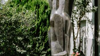 Moderne Figurenskulptur aus Bronze, stehend zwischen Blumen und Sträuchern in Vaduz – Werk von Herbert Albrecht.