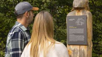 Zwei Personen lesen eine Infotafel mit Sage am WalserSagenWeg, auf einer geschnitzten Holzfigur