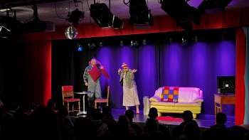 Kabarettszene im K-BUM in Liechtenstein – bunte Bühne mit Sofa, Schauspielerduo und Publikum im Saal.