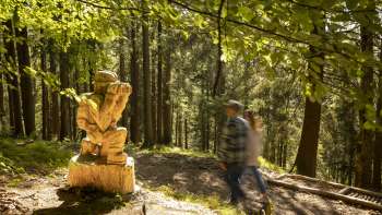 Wanderer passieren eine geschnitzte Figur mit Fernglas am WalserSagenWeg