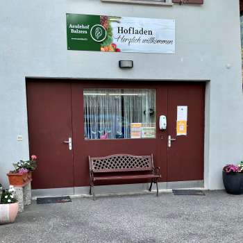 Eingang zum Hofladen Aeulehof mit rotem Tor, Sitzbank und Blumendekoration in Triesen, Liechtenstein.
