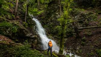 Wanderer mit Rucksack vor einem idyllischen Wasserfall bei Nendeln, eingebettet in dichten Wald und moosbedeckte Felsen.