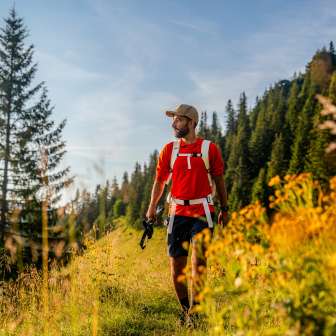 Wanderer mit rotem Shirt auf einem sonnigen Bergweg bei Malbun, umgeben von Wiesenblumen, Nadelwald und alpiner Kulisse in Liechtenstein