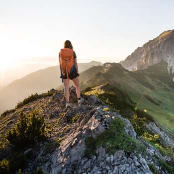 Wanderin mit Rucksack steht auf einem Bergkamm in Liechtenstein und blickt in die aufgehende Sonne