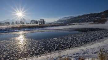 Sonnenaufgang am Egelsee in Mauren im Winter mit glitzernder Eisfläche, Schnee und sonniger Bergkulisse.