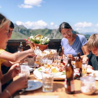 Familie beim gemeinsamen Mittagessen auf einer Sonnenterrasse in Sarreis, umgeben von Bergen und regionalen Spezialitäten