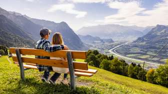 Panorama-Aussicht von Triesenberg über das Rheintal Paar sitzt auf einer Bank in Triesenberg und blickt auf das Rheintal in Liechtenstein mit beeindruckendem Alpenpanorama