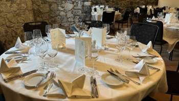 Elegant gedeckter runder Tisch mit weißen Tischdecken, Servietten und Speisekarten vor einer Steinwand.