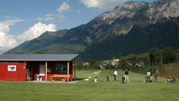 Golferinnen und Golfer auf der Driving Range in Liechtenstein mit Blick auf grüne Wiesen, ein rotes Clubhaus und die umliegende Berglandschaft bei blauem Himmel.