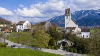 Panoramablick auf den Kirchhügel Bendern mit historischer Kirche, umgeben von gepflegten Wegen, Bäumen und traditioneller Architektur vor alpiner Bergkulisse.