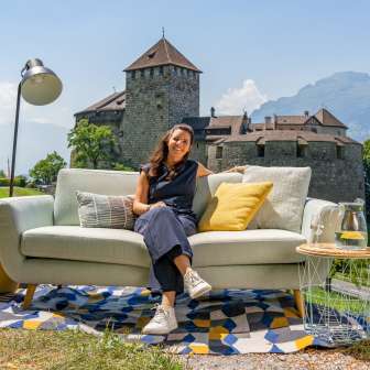 Frau sitzt auf einem Sofa mit Blick auf Schloss Vaduz und die Berge Liechtensteins, modernes Ambiente im Freien mit Dekoration