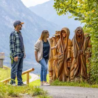 Paar betrachtet geschnitzte Holzfiguren am Walser Sagenweg in Triesenberg mit Blick ins Tal und Bergen im Hintergrund