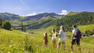 Eine Familie wandert durch eine blühende Sommerwiese in Malbun, Liechtenstein, und genießt den Blick auf die umliegenden Alpen.