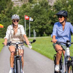 Zwei Frauen auf E-Bikes bei sommerlicher Tour mit Flaggen von Liechtenstein, Schweiz und Österreich im Hintergrund