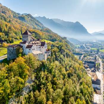Luftaufnahme von Schloss Vaduz auf einem Hügel über der Stadt, mit Alpenpanorama und Blick ins Rheintal bei klarer Sicht.