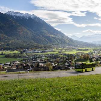 Grüner LIEmobil Bus fährt eine kurvige Straße oberhalb von Liechtenstein entlang mit Blick auf das Rheintal und die umliegenden Alpen