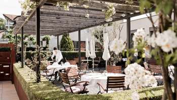 Eine elegante Terrasse mit weißen Tischen, gedeckt mit Servietten und Gläsern unter einem Holzpavillon, umgeben von üppigem Grün.
