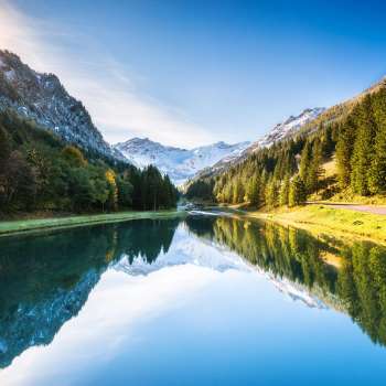 Gänglesee in Steg mit Bergspiegelung, umgeben von Wald, Sonnenstrahlen über dem Hang.