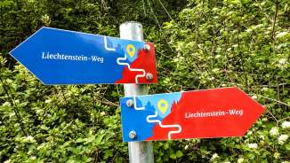 Bunter Wegweiser des Liechtenstein-Wegs mit roten und blauen Pfeilen vor grünem Waldhintergrund