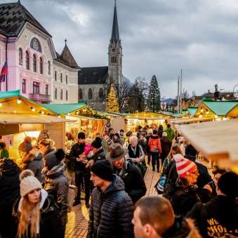 Stimmungsvolle Abendaufnahme des Weihnachtsmarkts in Vaduz 2018 mit festlich geschmückten Marktständen und Menschenmenge vor Regierungsgebäude und Kathedrale