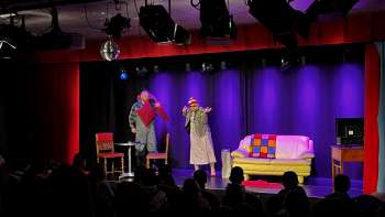 Kabarettszene im K-BUM in Liechtenstein – bunte Bühne mit Sofa, Schauspielerduo und Publikum im Saal. Kabarettszene im K-BUM in Liechtenstein – bunte Bühne mit Sofa, Schauspielerduo und Publikum im Saal.