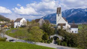 Panoramablick auf den Kirchhügel Bendern mit historischer Kirche, umgeben von gepflegten Wegen, Bäumen und traditioneller Architektur vor alpiner Bergkulisse.