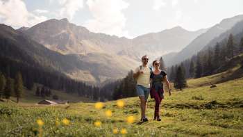 Zwei Personen wandern durch eine weite Alpwiese mit Blick auf die umliegenden Berge der Alp Valüna bei sonnigem Wetter