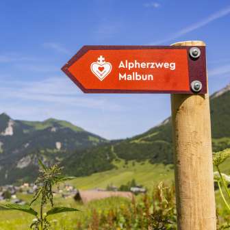 Rotes Schild mit der Aufschrift „Alpherzweg Malbun“ vor malerischer Bergkulisse und Sommerwiese in Liechtenstein.